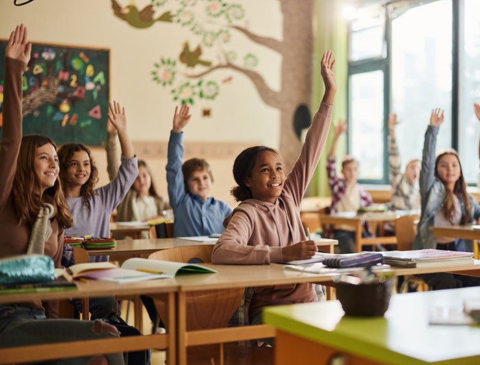 elementary school children sit in classroom with hands raised to answer questions