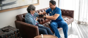 National Rural Health Day Age Friendly Image - A visiting nurse sitting on sofa with a elderly woman taking her blood pressure in her home.
