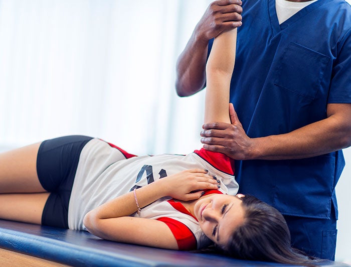 A therapist in scrubs treats a teen female athlete, stretching her arm