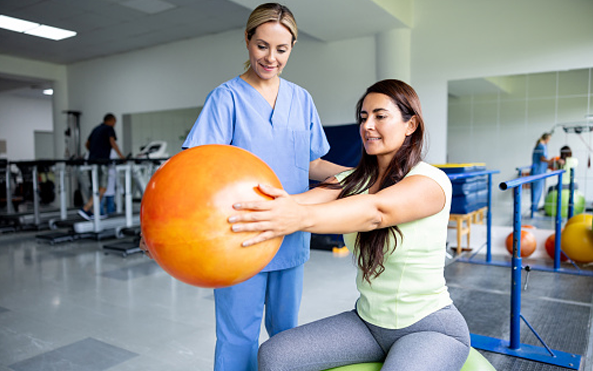 9 Inpatient Rehab Hospital Best Practices. A physical therapist works with a patient who is sitting on a large exercise ball while holding a small exercise ball straight out in front of her.
