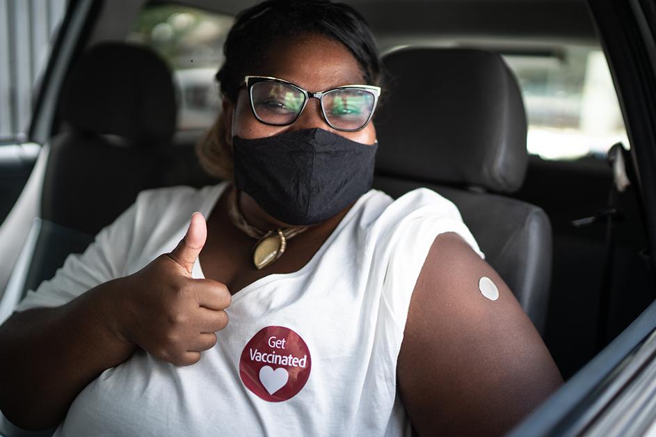 COVID-19 and Vaccination Efforts. A woman wearing a mask with a Get Vaccinated sticker on her shirt shows her vaccine location Band-Aid.