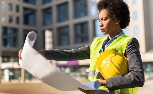Cleveland Clinic, University Hospitals DEI Supplier Accelerator Program Aims to Level the Playing Field. A construction worker in reflective vest holding a hard hat safety helmet under her arm looks at blueprints she is holding unfurled in her hands.