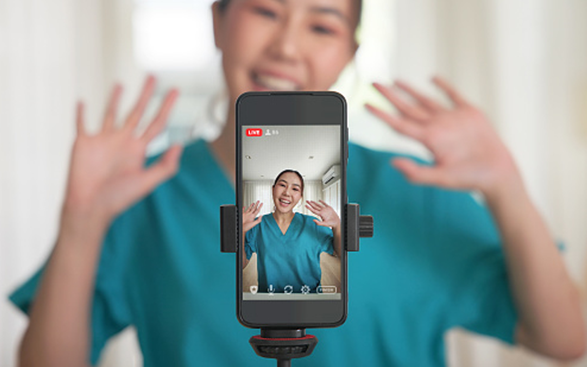 How Health Systems Can Retain Gen Z Workers. A nurse stands in front of a mobile phone that is recording a video of her.