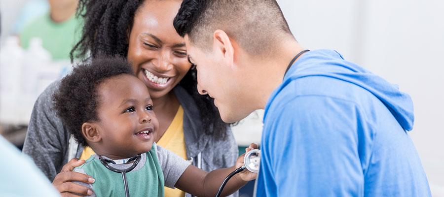 Health Equity. Latino doctor examining African-American child on mother's lap who is playing with the doctor's stethoscope.