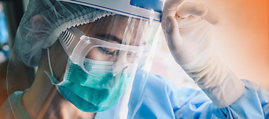 How to Help Clinicians Stay Resilient during COVID-19. A health care working in gown, gloves, surgical mask, goggles, surgical cap, and face shield with head down and the back of her hand on her forehead.