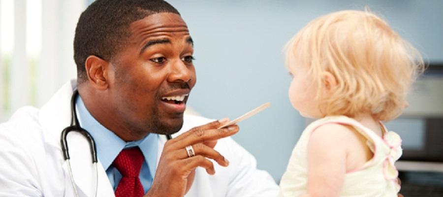 A Black male doctor with a tongue depressor in his right hand examines a small child. 