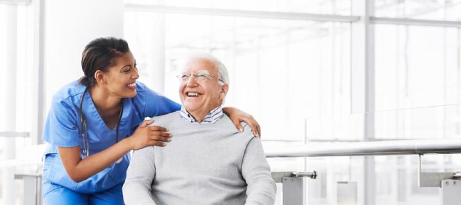 A smiling, female health care worker wearing scrubs stands next to a smiling, elderly male patient, with her arm around his shoulder.