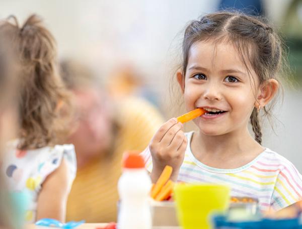 A smiling young girl sits at a school dining table eating a healthy meal