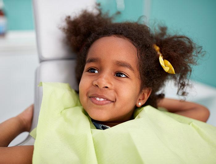 A young girl with afro puffs and yellow bows in her hair sits smiling in a dentist's chair