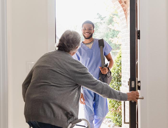 A smiling, young, male caregiver in scrubs is greeted at the front door by his elderly patient