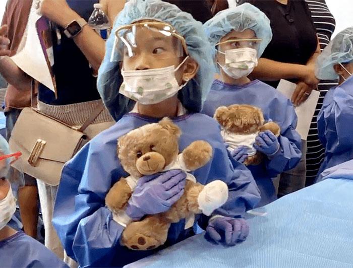 Pediatric patients in PPE holding teddy bears sit at a table surrounded by standing adults