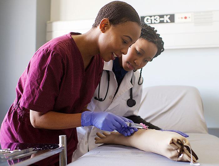A female health worker in scrubs practices needle insertion in a training arm while another health worker in a white coat looks on