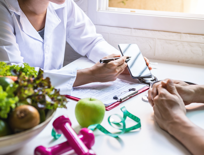 A physician counsels a patient on diet and exercise, indicated by a salad and dumbbells on her desk
