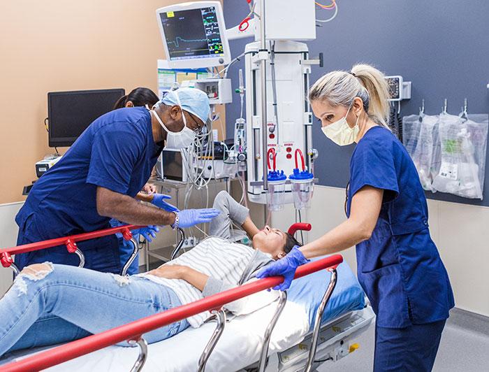 Two emergency department health workers in scrubs treat a female patient on a hospital bed in a triage bay