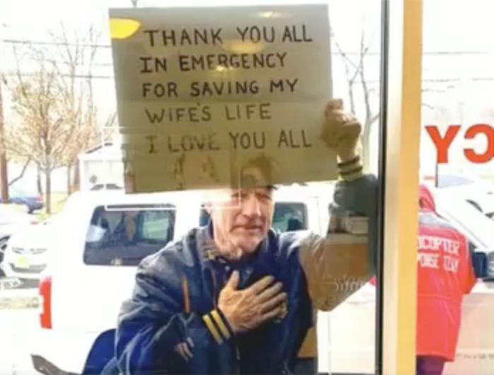 A man stands outside an emergency room window holds a sign reading: Thank you all in Emergency for saving my wife's life. I love you all.