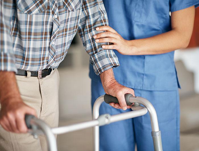 An aide in scrubs helps a patient stand with a walker