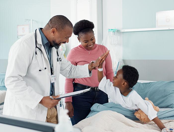 pediatric patient in hospital bed high-fives his doctor as his mother looks on